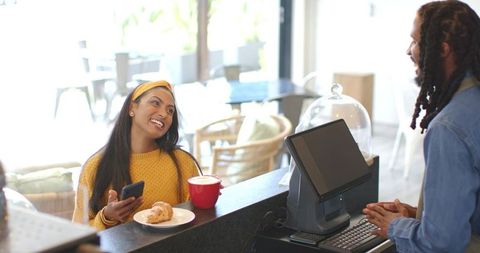 Indian woman smiling while ordering coffee and croissant at sunlit cafe counter
