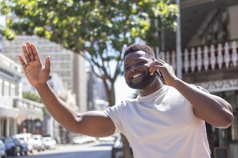 Smiling african man walking downtown, talking on smartphone, waving under sunlit tree canopy