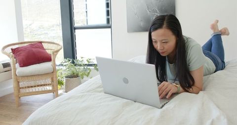 Asian woman working remotely on laptop lying on bed in bright minimalist home office