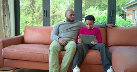 African American Father and Son Sharing Tablet on Leather Sofa by Garden Window Sunlight