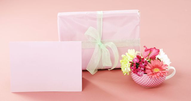 A gift wrapped in pink paper with a green ribbon is accompanied by a bouquet of colorful flowers in a polka-dotted cup and a blank pink card, with copy space. Ideal for occasions like Mother's Day or Valentine's Day, the setup conveys a thoughtful gesture of affection and celebration.