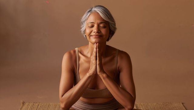 Senior woman practicing meditation and prayer pose on woven mat in beige wellness studio