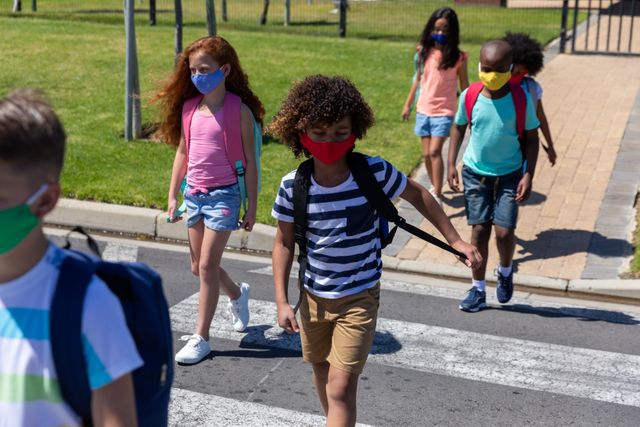 Children wearing face masks and backpacks crossing the road on their way to school. This image can be used for topics related to education, health and safety, pandemic measures, back to school campaigns, and social distancing practices.