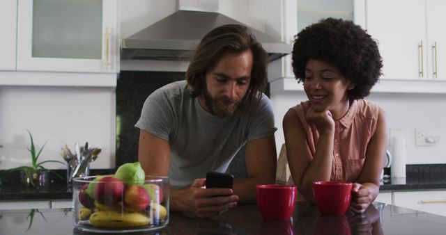 Biracial couple holding coffee cup using smartphone in the kitchen at home. staying at home in self isolation in quarantine lockdown