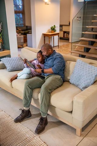 African american father and daughter sharing tablet on sofa, teaching and bonding