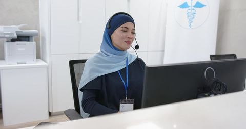 Smiling receptionist wearing light blue hijab answering calls and typing at clinic desk