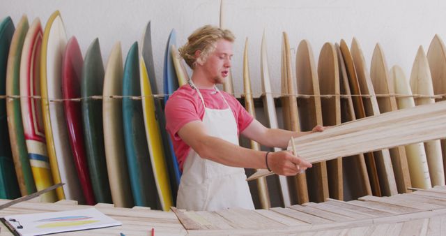 Young male surfboard maker in workshop carefully inspecting wooden frame while standing in front of various colored surfboards. Suitable for content related to craftsmanship, handmade goods, surfing, hobbyist woodworkers, and workshop activities.