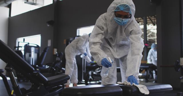 Healthcare professionals in full protective gear are seen disinfecting gym equipment with cleaning solution and wipes. This image is suitable for content related to COVID-19 sanitation measures, gym safety during the pandemic, health and hygiene practices, and public health protocols. It can be used in articles, blog posts, health and safety guidelines, and promotional materials for gyms, fitness centers, or health organizations.