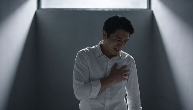 Asian man clutching chest experiencing acute pain in dim room with dramatic window light