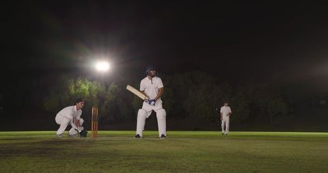 Diverse male cricketers batting under floodlights at night on pitch, helmeted batsman