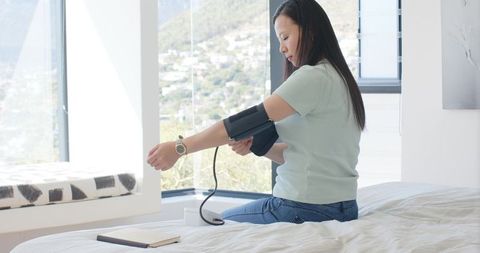 Asian woman measuring blood pressure on bed in bright modern bedroom
