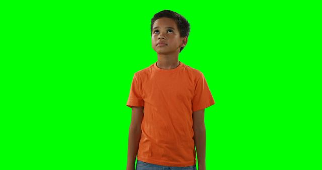 Boy in an orange shirt looking up against a green screen background. Can be used for visual effects, video editing, educational materials, or advertisements focusing on children's curiosity and thinking.