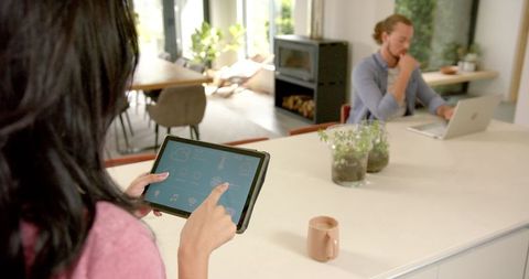 Young Woman Managing Smart Home App on Tablet While Partner Works Remotely