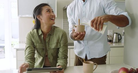 Midlife asian couple enjoying morning at kitchen island, man pouring milk, woman using tablet