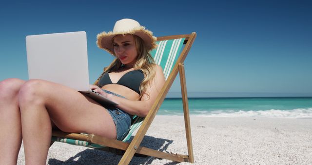 This image features a young woman in a straw hat sitting on a deckchair on a sandy beach, working on a laptop. The clear blue sky and vibrant ocean waters in the background highlight a summer vacation setting. This can be used in articles or advertisements related to remote work, freelance opportunities, summer vacations, digital nomads, and technology.