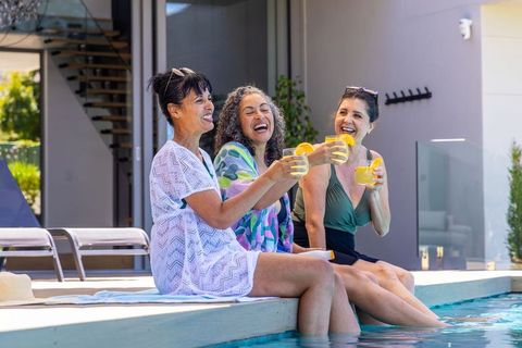 Diverse women laughing and toasting with citrus drinks by poolside on modern patio