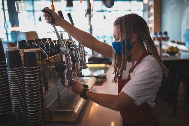 Barista with dreadlocks wearing face mask and apron preparing coffee in a cafe. Ideal for content related to small businesses, coffee culture, pandemic safety measures, and professional barista training. Useful for articles, blogs, and marketing materials promoting cafes, coffee shops, and hygiene practices during COVID-19.