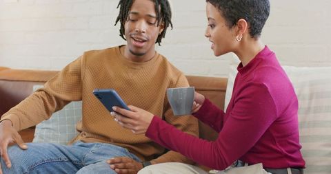 Couple sharing smartphone on cozy sofa while woman holds mug casual home lifestyle
