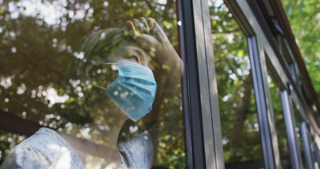 Woman wearing face mask and looking through window with tired expression. Reflection of outdoors and nature visible on glass. Perfect for themes of health, safety, isolation, pandemic, and quarantine.