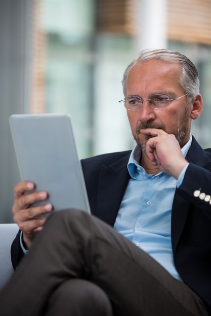 Senior businessman sitting in modern office, holding digital tablet, and looking thoughtful. Ideal for use in business, technology, and corporate settings to depict professionalism, concentration, and modern work environments.