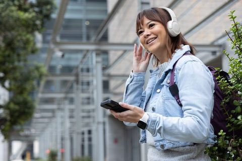 Asian woman enjoying music with white headphones while using smartphone in modern urban corridor