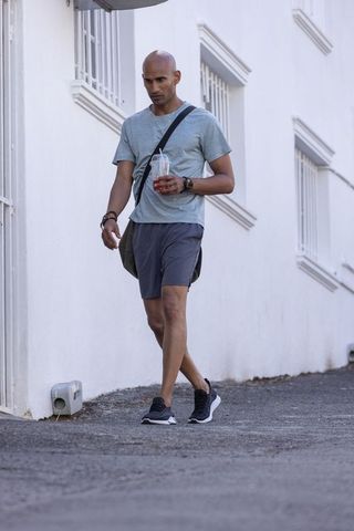 African man walking with iced drink and crossbody bag along white stucco urban alleyway