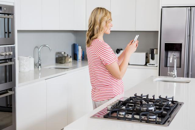 Blonde woman standing in modern kitchen, casually using mobile phone. Ideal for conveying themes of home life, technology use in daily life, modern kitchen design, or domestic chores. Suitable for advertisements, blogs, and articles related to home decor, lifestyle, and technology.