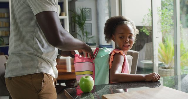 An African American father helps his young daughter get ready for school by placing items into her backpack. They are in a modern kitchen with natural light streaming in. This image is perfect for themes related to family bonding, morning routines, parent-child relationships, and education.