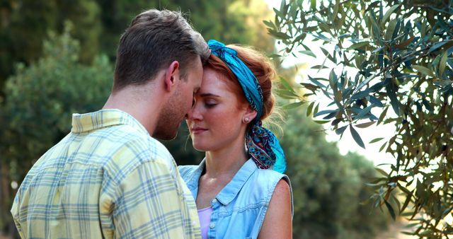 A young Caucasian couple shares an intimate moment, with the woman wearing a blue headscarf and the man in a checkered shirt, surrounded by greenery, with copy space. Their affectionate embrace suggests a romantic connection in a serene outdoor setting.