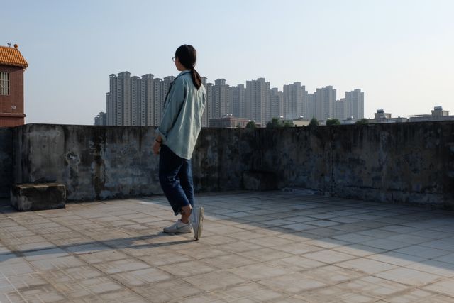 Woman stands on a rooftop, gazing at city high-rises in the distance under a clear sky. Suitable for urban lifestyle, solitude, or architecture themes.