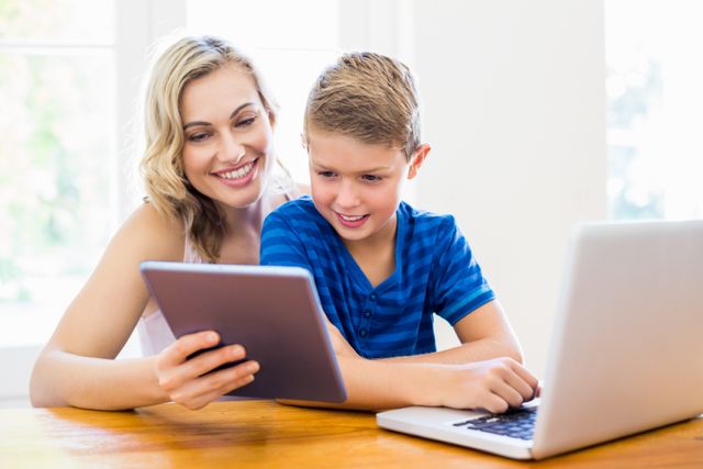 Mother and son enjoying time together using a laptop and digital tablet at home. Perfect for illustrating family bonding, modern parenting, technology in education, and home learning environments.