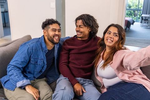 Friends laughing and taking selfie while lounging on gray sofa in bright modern apartment
