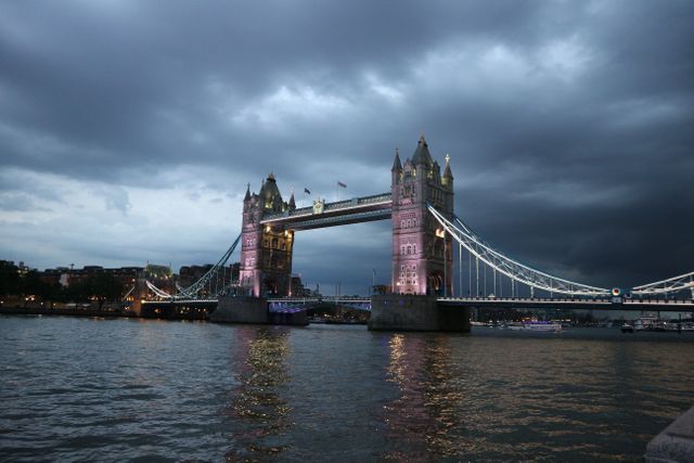 Tower Bridge is illuminated at dusk with a dramatic cloudy sky in this scenic view. Ideal for use in travel and tourism articles, cityscape photography collections, historical architecture features, and promotional materials highlighting London as a travel destination.