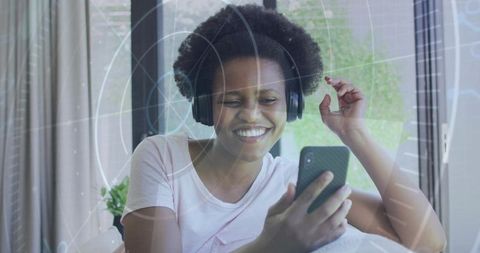 Smiling woman listening to music with over-ear headphones using smartphone at home