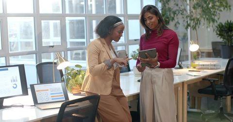 Two diverse female coworkers collaborating and discussing project on tablet in sunlit office