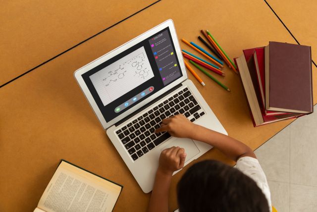 Child engaged in online learning at home, notes on laptop, textbooks and pencils on table. Ideal for concepts related to homeschooling, e-learning platforms, virtual classrooms, technology in education, elementary education, and distance learning.