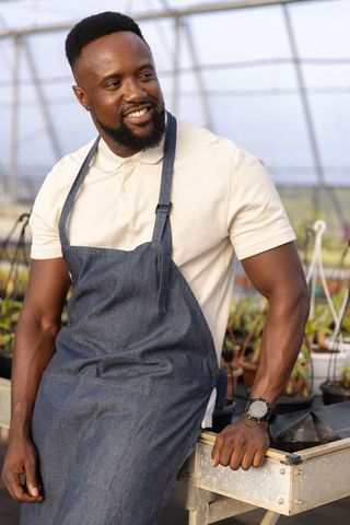 Smiling gardener leaning in greenhouse wearing denim apron and checking seedling trays