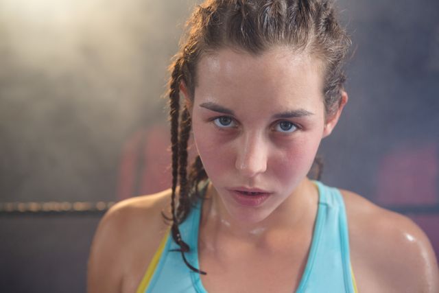 Young female boxer with braided hair and sweaty face showing determination and focus in a fitness studio. Ideal for use in articles, advertisements, and promotions related to fitness, sports training, women's empowerment, and athleticism.