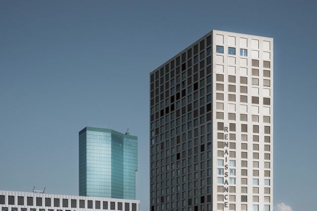 Image features a modern cityscape with contemporary skyscrapers under a clear blue sky. The foreground highlights a high-rise building with grid-like windows and the word 'RENAISSANCE' visibly written on its side. The background includes another tall, glass-covered structure. Ideal for use in business, real estate, and urban planning contexts.