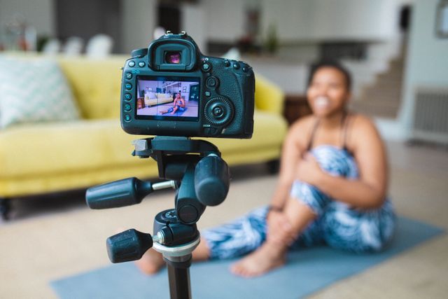 Young African American woman recording a fitness video while sitting on a yoga mat in her home. Perfect for use in articles about fitness routines, healthy lifestyle tips, social media influence, or home workout guides.