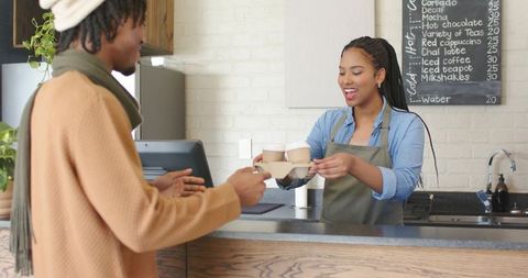 Smiling diverse barista handing two takeaway coffees to customer at urban cafe counter