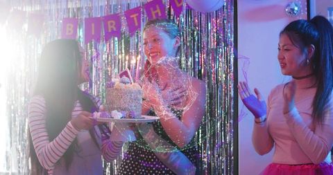 Young women celebrating birthday with candlelit sprinkle cake and disco tinsel backdrop