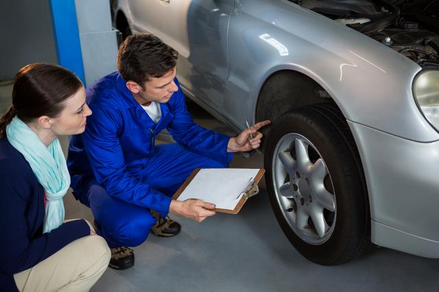 Mechanic in blue overalls showing car issue to customer in repair garage. Mechanic holding clipboard, pointing at car tire. Customer attentively listening. Useful for automotive service promotions, customer service training materials, and repair shop advertisements.