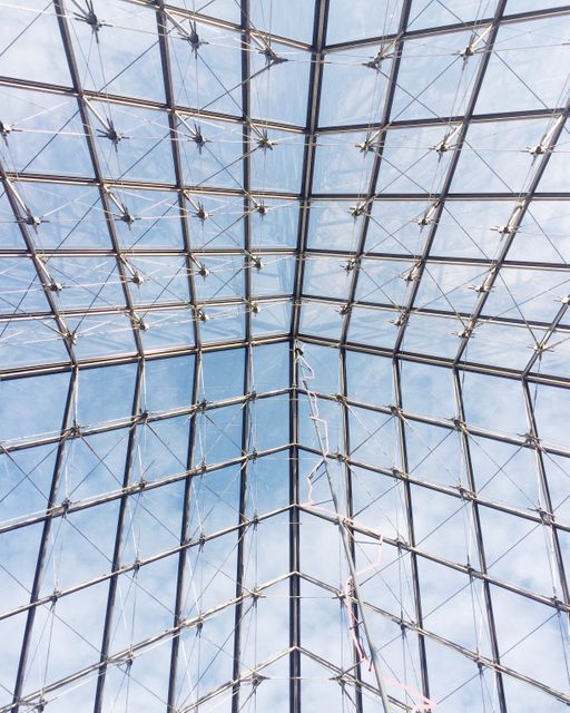Showing a modern glass roof architecture featuring a sturdy metal framework, this photo captures the intricate geometric design against a clear sky backdrop. Ideal for usage in architectural reviews, modern construction promotions, and illustrating articles about innovative building designs and urban development.
