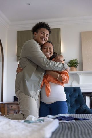 Multiracial couple embracing in cozy bedroom while preparing nursery and baby clothes