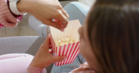 Friends sharing popcorn on couch in pastel sweaters cozy home movie night snack moment
