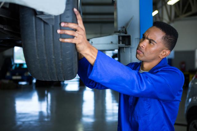 Mechanic in blue coveralls inspecting and fixing a car tire in a repair garage. Ideal for use in automotive service promotions, repair and maintenance guides, mechanical engineering materials, and auto service center advertisements.