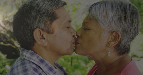 Kissing senior couple sharing tender moment in sunlit garden with green bokeh romantic affection