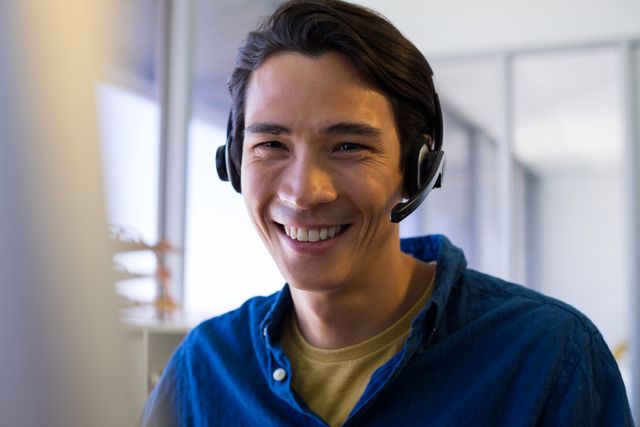 This image depicts a young male executive wearing a headset and smiling at his desk in an office environment. It is ideal for use in business, customer service, and communication-related content. The image can be used to represent professionalism, support, and a positive work atmosphere in corporate settings.