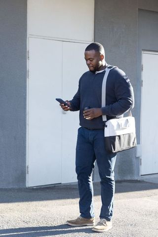 African american commuter checking smartphone while carrying coffee and messenger bag